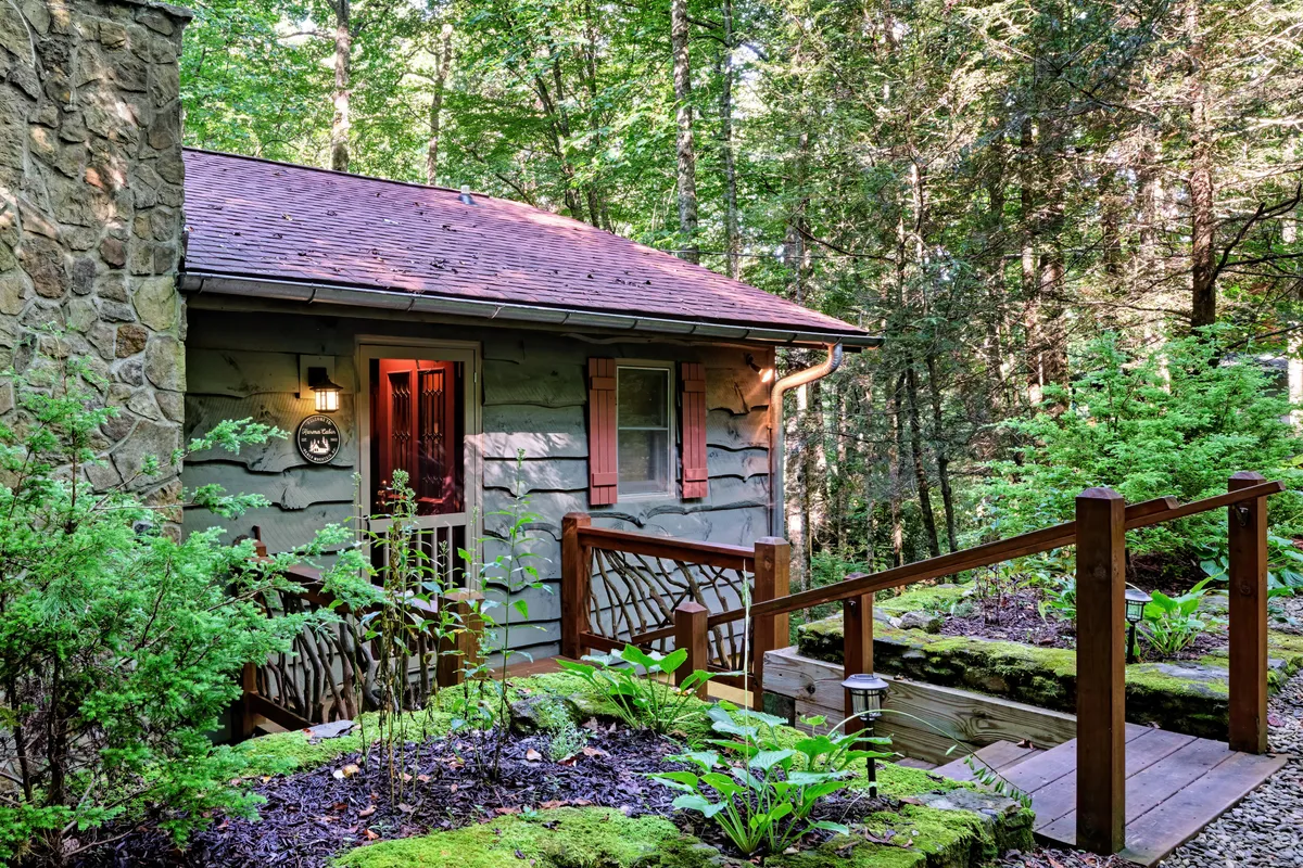A charming stone cabin sits nestled among lush greenery, with a wooden walkway inviting guests to relax on the porch or explore the surrounding nature trails. The peaceful setting suggests a perfect spot for outdoor gatherings, barbecues, or morning coffee with friends.