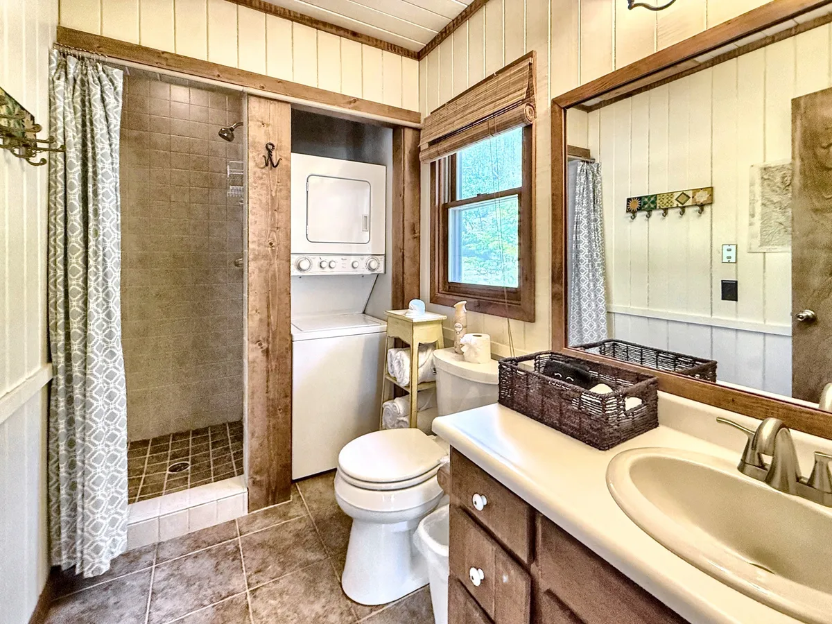 A bright, practical bathroom and laundry area featuring a glass-enclosed shower, efficient laundry units, a sink with vanity, and a sunlit window. The overall scene feels clean, organized, and functional—ideal for the needs of busy families.
