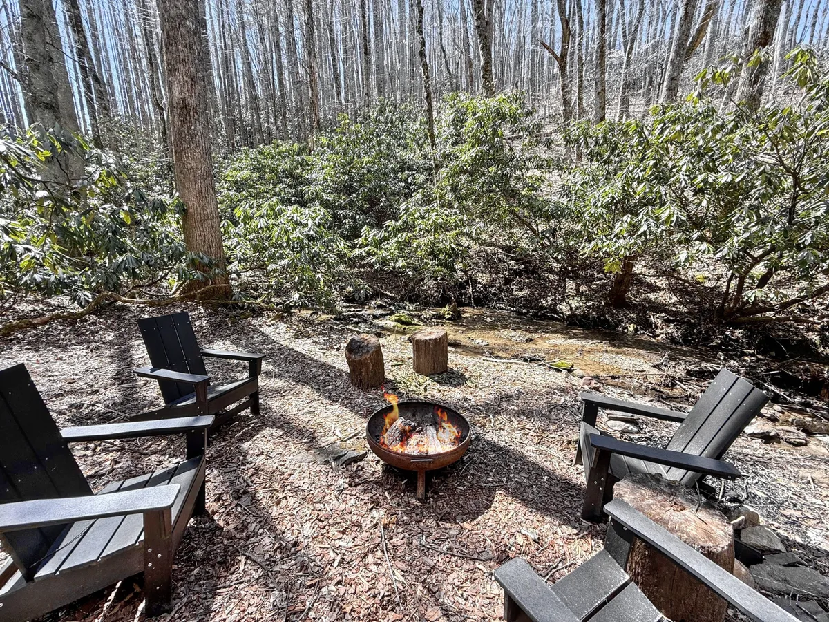 An inviting outdoor fire pit area with wooden chairs set beside a gently flowing stream. The scene is surrounded by nature, with the soft light of sunset creating a relaxed mood ideal for cozy campfire gatherings or stargazing.