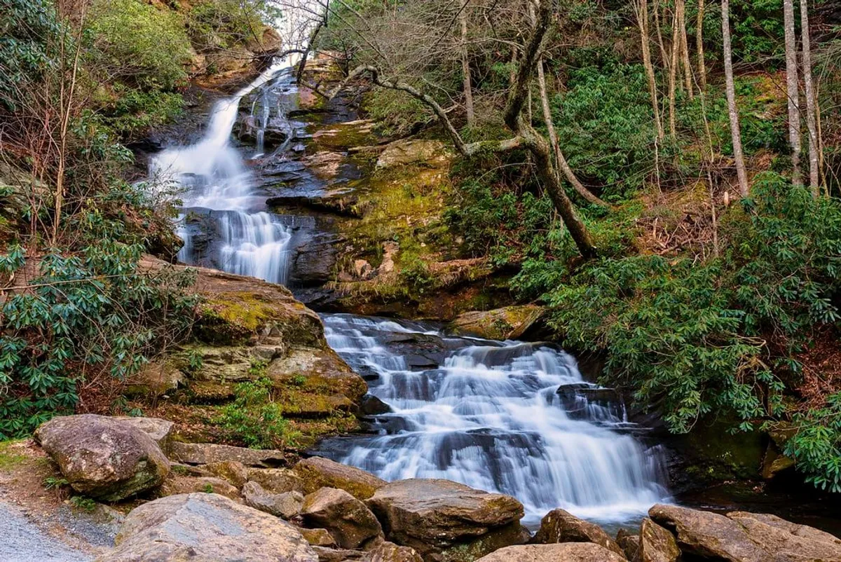 A scenic waterfall cascades down rocks, surrounded by vibrant greenery and foliage. The lush setting invites visitors to explore, hike, or simply take in the tranquil beauty of the natural landscape. Sunlight filters through the trees, creating dappled patterns on the rocks and water.