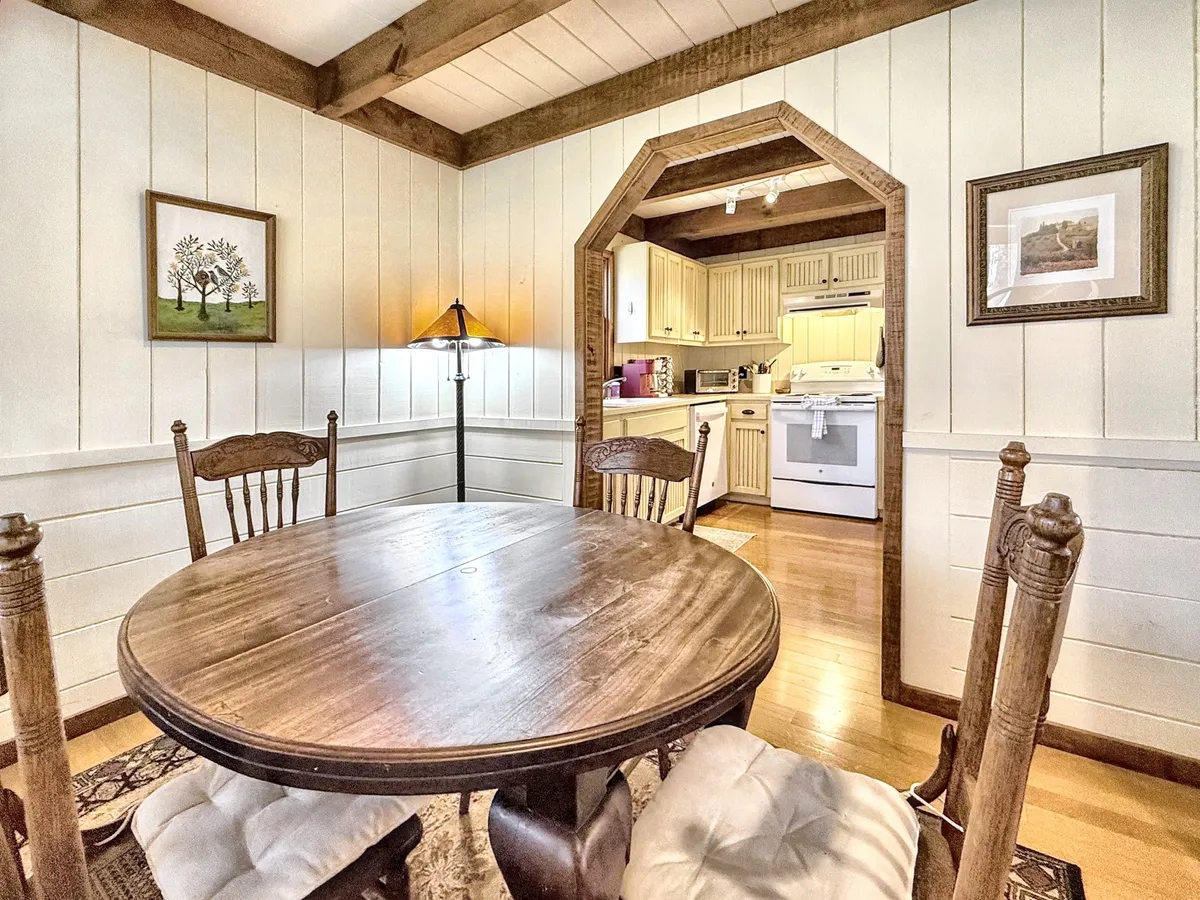 A rustic wooden dining table set beside an arched doorway to the kitchen, with welcoming wall art and soft lighting from a floor lamp. The space is designed for dining, group conversations, family gatherings, and spirited game nights, all in a warm and homey atmosphere.