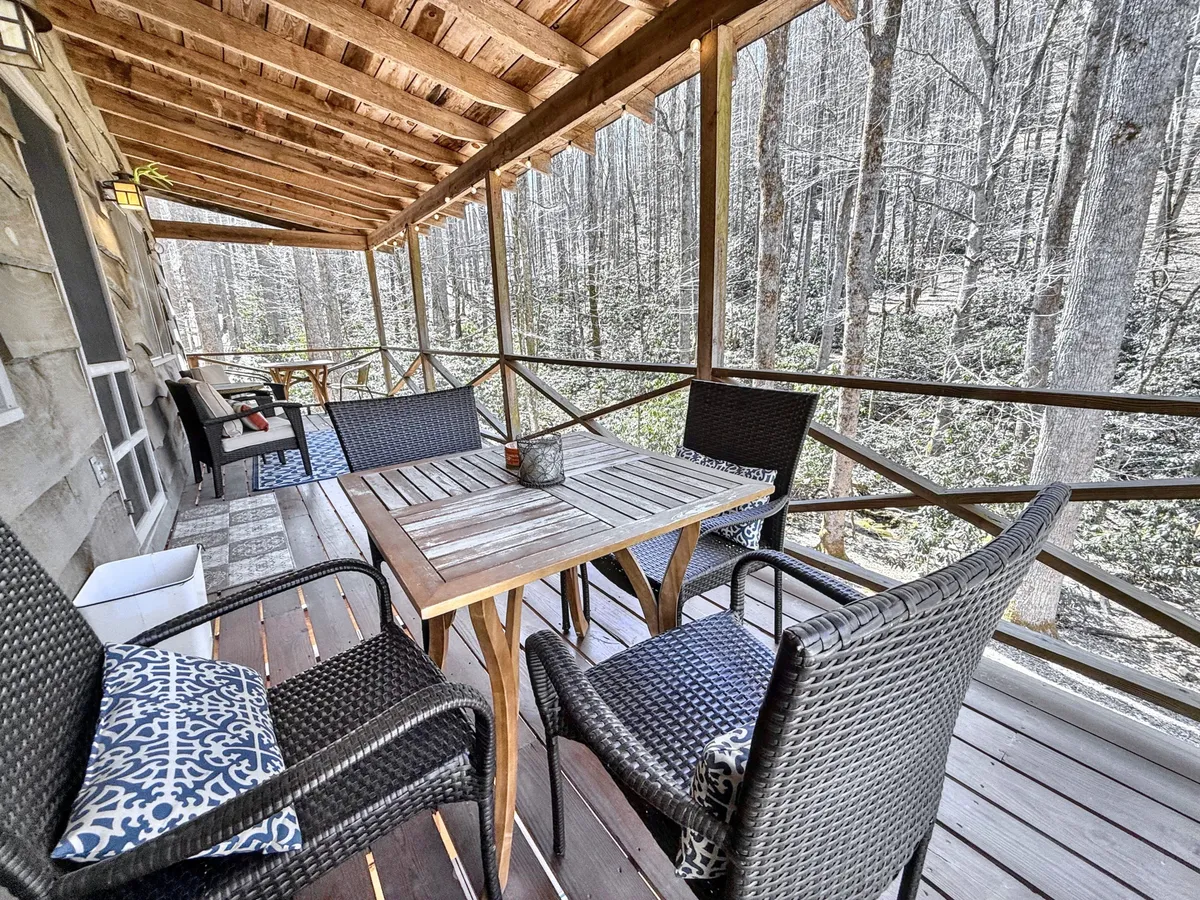 A serene exterior scene featuring a wooden table and woven chairs set on a covered deck, surrounded by a lush forest. The table is positioned to enjoy sunset views through the trees, and decorative pillows add a cozy touch. The setting is perfect for enjoying morning coffee, reading, or relaxing outdoors.