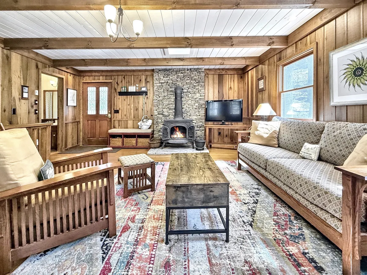 The image showcases a warm and inviting cabin interior, centered around a rustic stone fireplace. Cozy wooden furniture surrounds the hearth, accented by a vibrant, colorful rug, creating a perfect space for relaxing, reading by the fire, or enjoying quality time with family and friends.