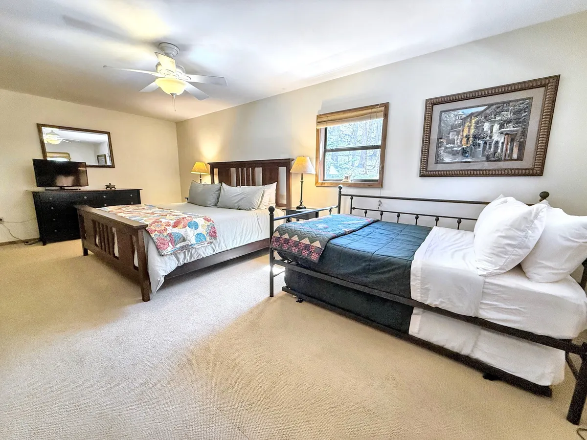 A cozy bedroom featuring a queen bed adorned with a colorful quilt, a twin bed with a dark blue throw, warm wooden furniture, framed artwork on the wall, and softly glowing table lamps. The arrangement suggests space for both relaxation and quiet conversation, with inviting textures and restful lighting.